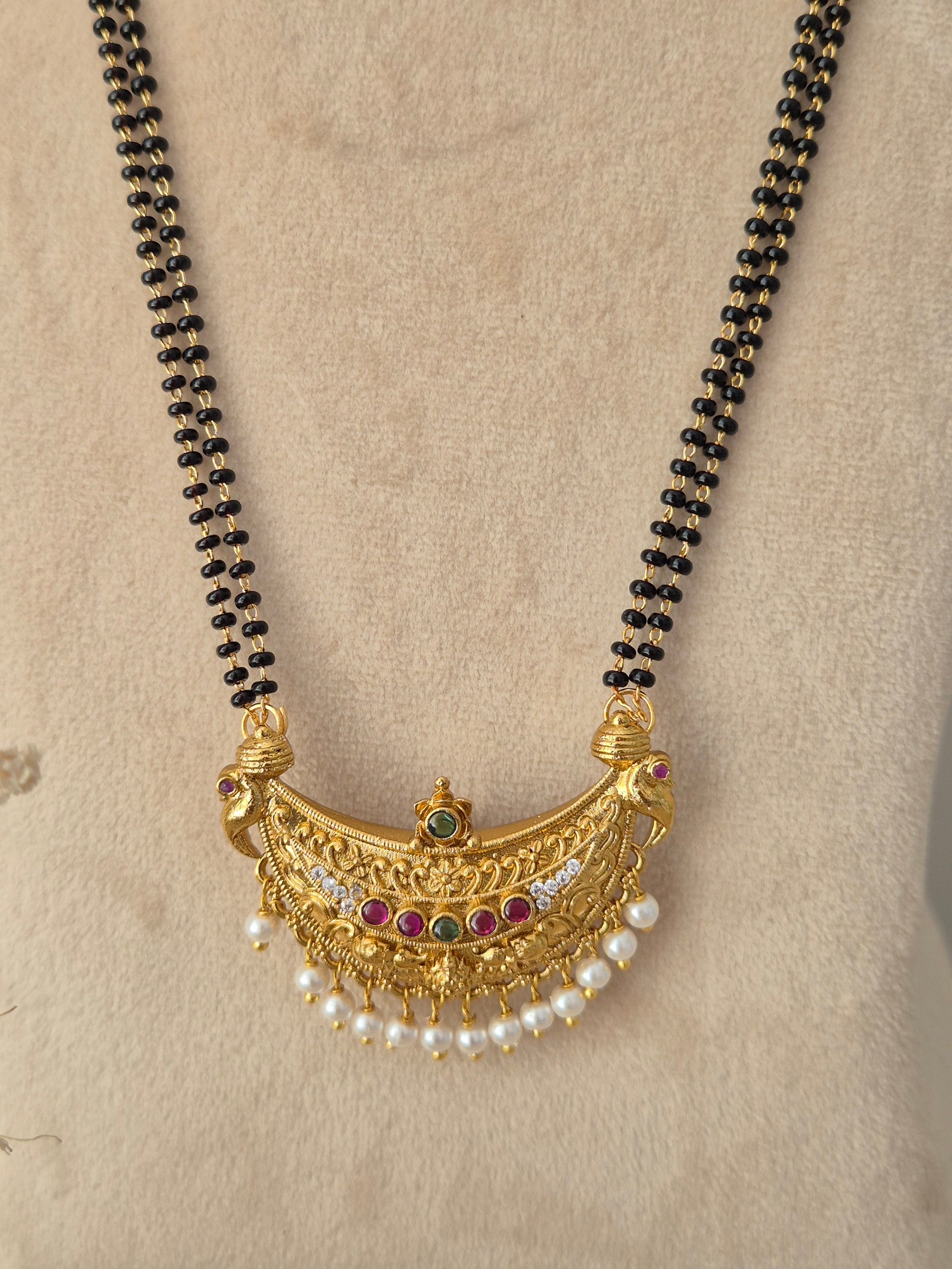 Close-up view of the Janani Mangalsutra crescent gold pendant with red and green stones, pearl drops, and black-beaded chain