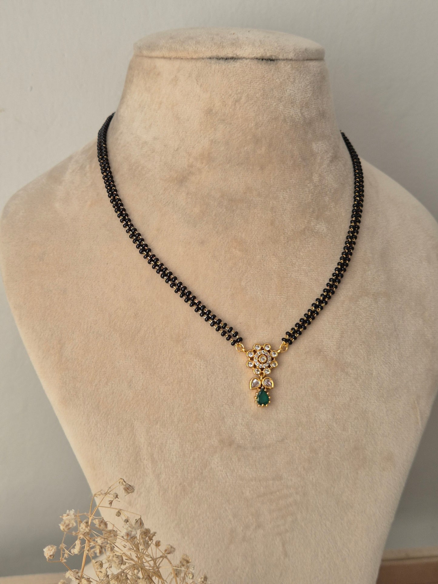 Balaka Mangalsutra featuring a fine double-strand black bead chain connected to a delicate gold floral motif pendant studded with clear CZ stones and finished with a green teardrop stone drop