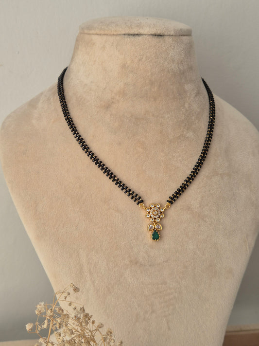 Balaka Mangalsutra featuring a fine double-strand black bead chain connected to a delicate gold floral motif pendant studded with clear CZ stones and finished with a green teardrop stone drop