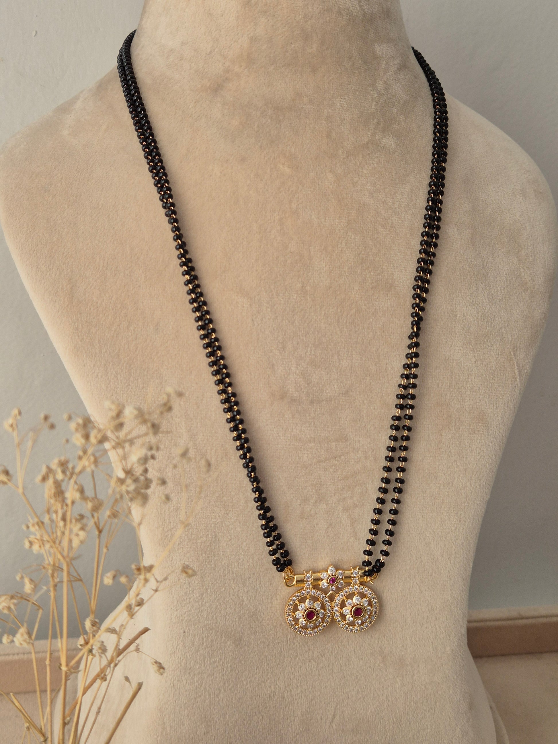 Baani Mangalsutra featuring a long double-strand black bead chain connected to twin round gold pendants embellished with clear stones and a central red gemstone