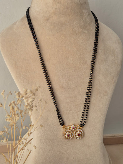 Baani Mangalsutra featuring a long double-strand black bead chain connected to twin round gold pendants embellished with clear stones and a central red gemstone