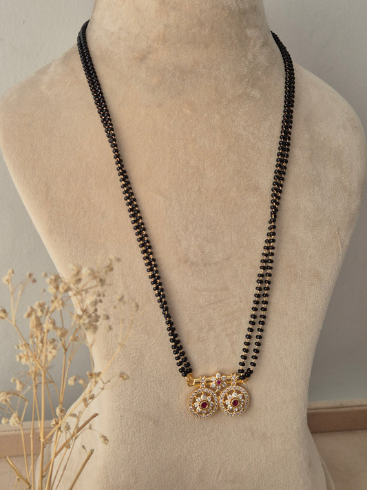 Baani Mangalsutra featuring a long double-strand black bead chain connected to twin round gold pendants embellished with clear stones and a central red gemstone