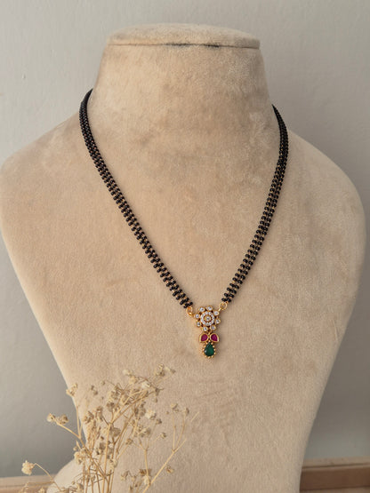 Anjika Mangalsutra featuring a fine double-strand black bead chain connected to a small gold floral pendant set with clear CZ stones, finished with red stone accents and a green teardrop drop