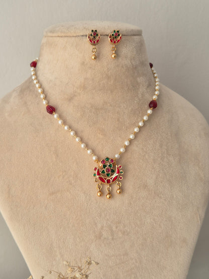 Aloka Necklace Set with a white pearl and red bead chain attached to a gold pendant studded with red and green stones and hanging gold beads, paired with matching stud drop earrings