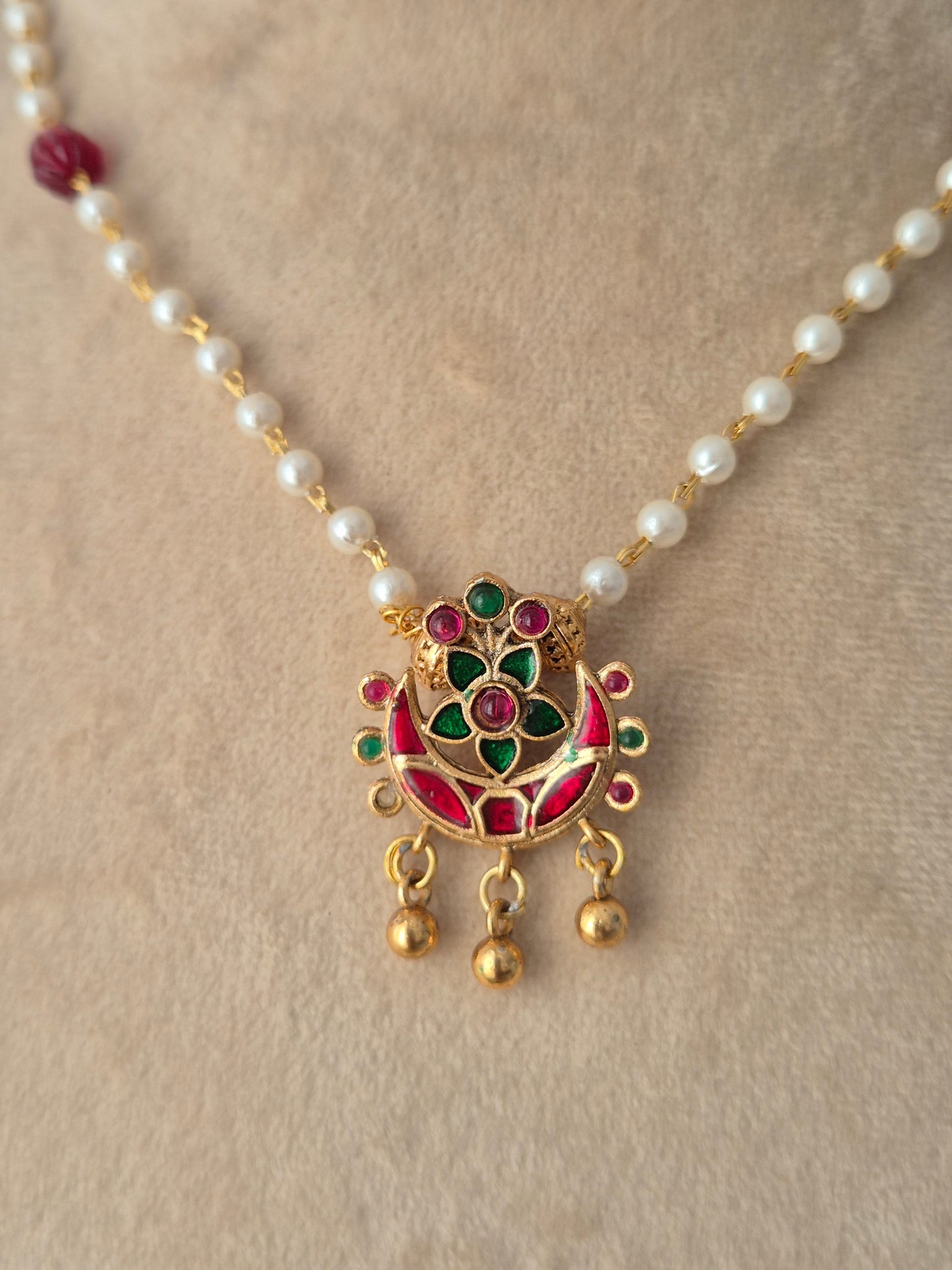 Close-up view of the Aloka Necklace gold crescent pendant with red and green stones, gold bead drops, and white pearl chain