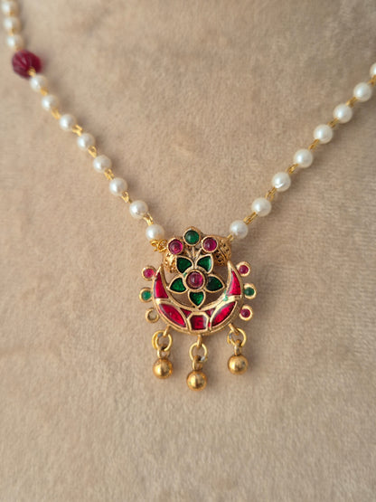 Close-up view of the Aloka Necklace gold crescent pendant with red and green stones, gold bead drops, and white pearl chain