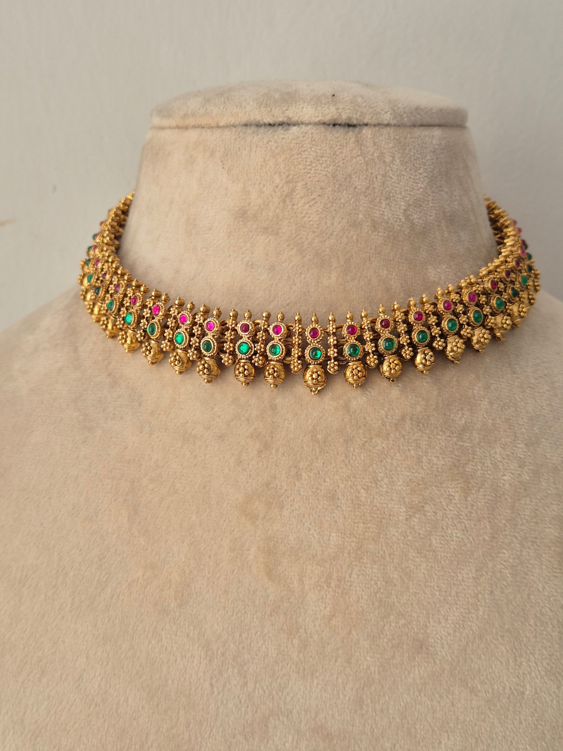 Traditional Alpana Choker Necklace Set crafted in antique gold-toned alloy, featuring a slim choker studded with alternating pink and green gemstones and dangling gold bead clusters, displayed alongside matching floral-topped jhumka earrings