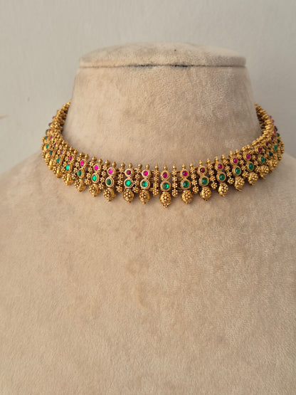 Traditional Alpana Choker Necklace Set crafted in antique gold-toned alloy, featuring a slim choker studded with alternating pink and green gemstones and dangling gold bead clusters, displayed alongside matching floral-topped jhumka earrings