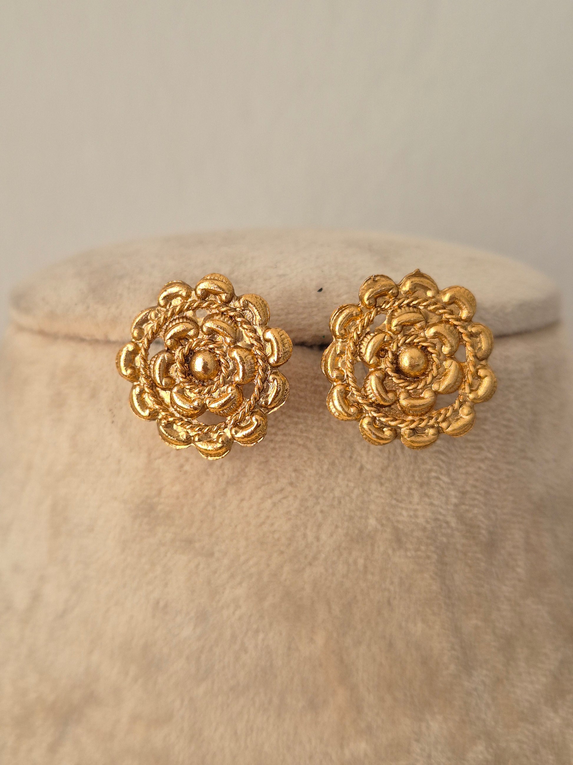 Close-up of a pair of gold-toned circular stud earrings featuring an intricate multi-layered floral design with textured rope-style borders
