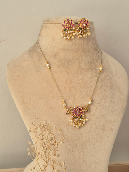 Ludhika Necklace Set showcasing a delicate gold chain necklace with a central lotus floral pendant studded with pink and green stones and small white pearl drops, displayed on a beige mannequin bust alongside matching floral stud earrings