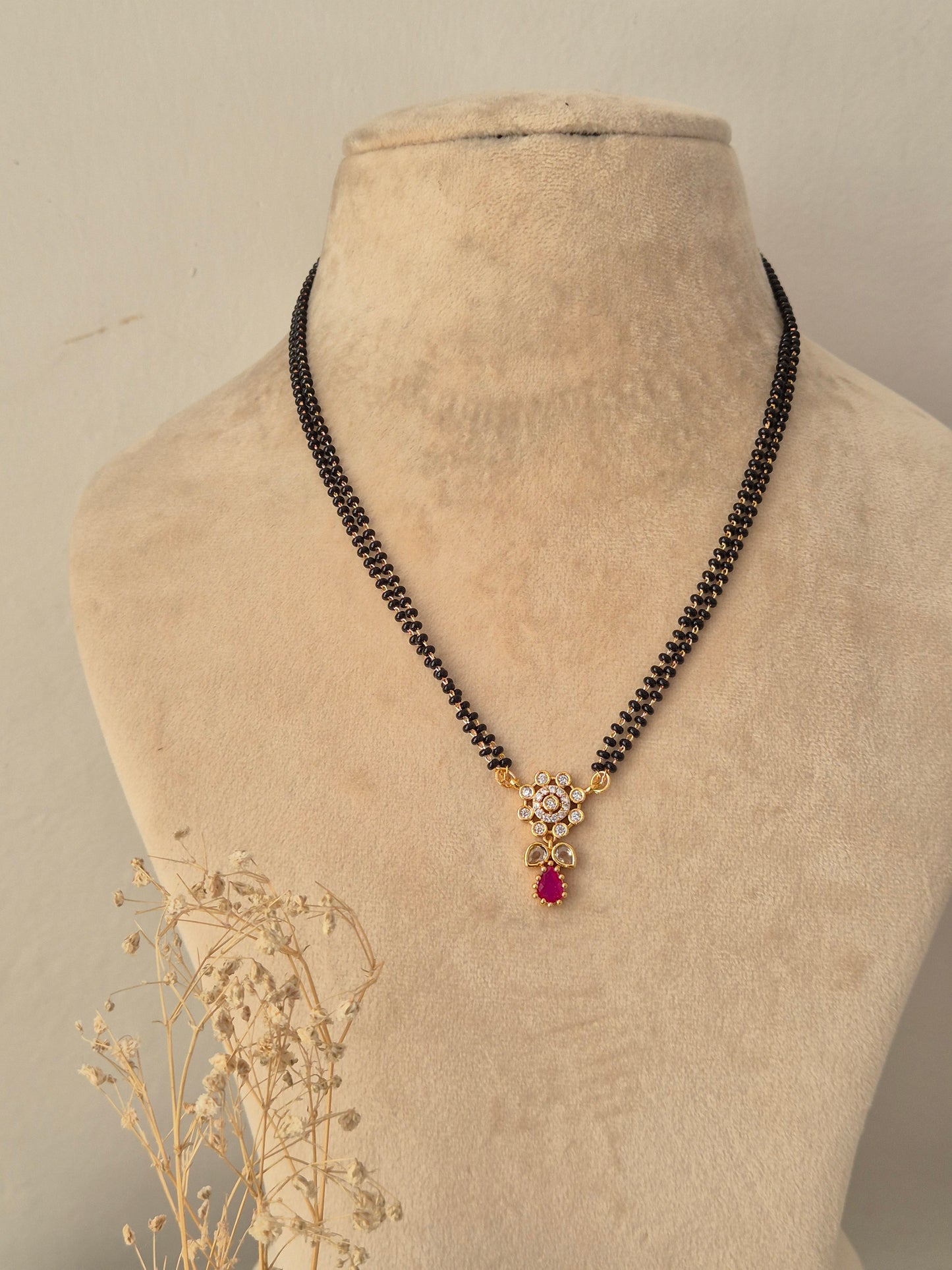 Sitika Mangalsutra showcasing a double-strand black bead chain with a small gold floral pendant embellished with clear CZ stones and a dangling magenta-pink teardrop stone