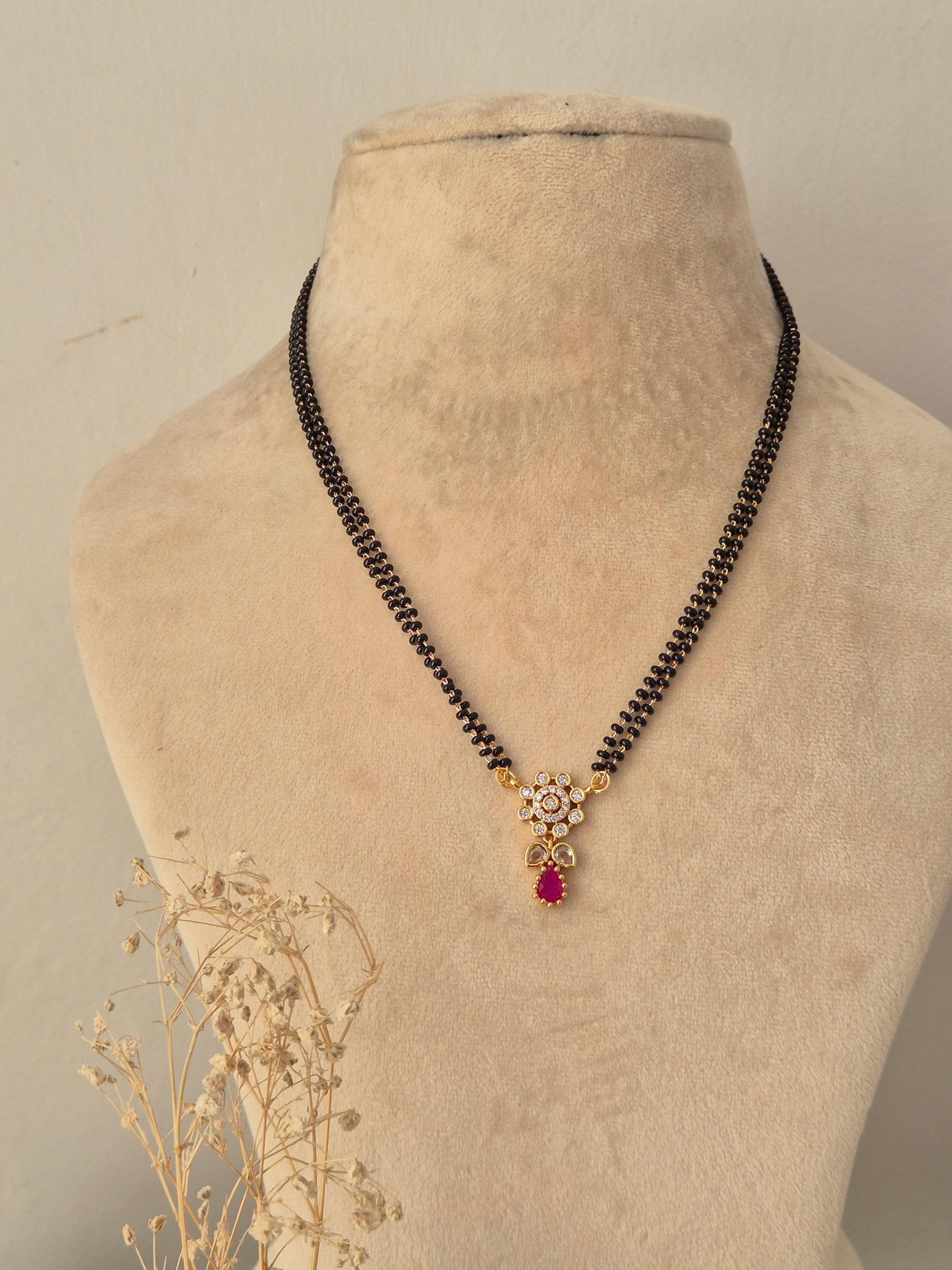 Sitika Mangalsutra showcasing a double-strand black bead chain with a small gold floral pendant embellished with clear CZ stones and a dangling magenta-pink teardrop stone