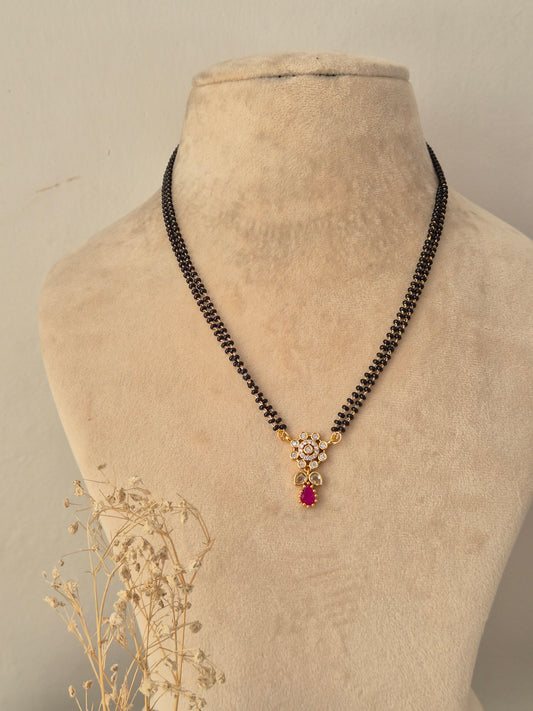 Sitika Mangalsutra showcasing a double-strand black bead chain with a small gold floral pendant embellished with clear CZ stones and a dangling magenta-pink teardrop stone