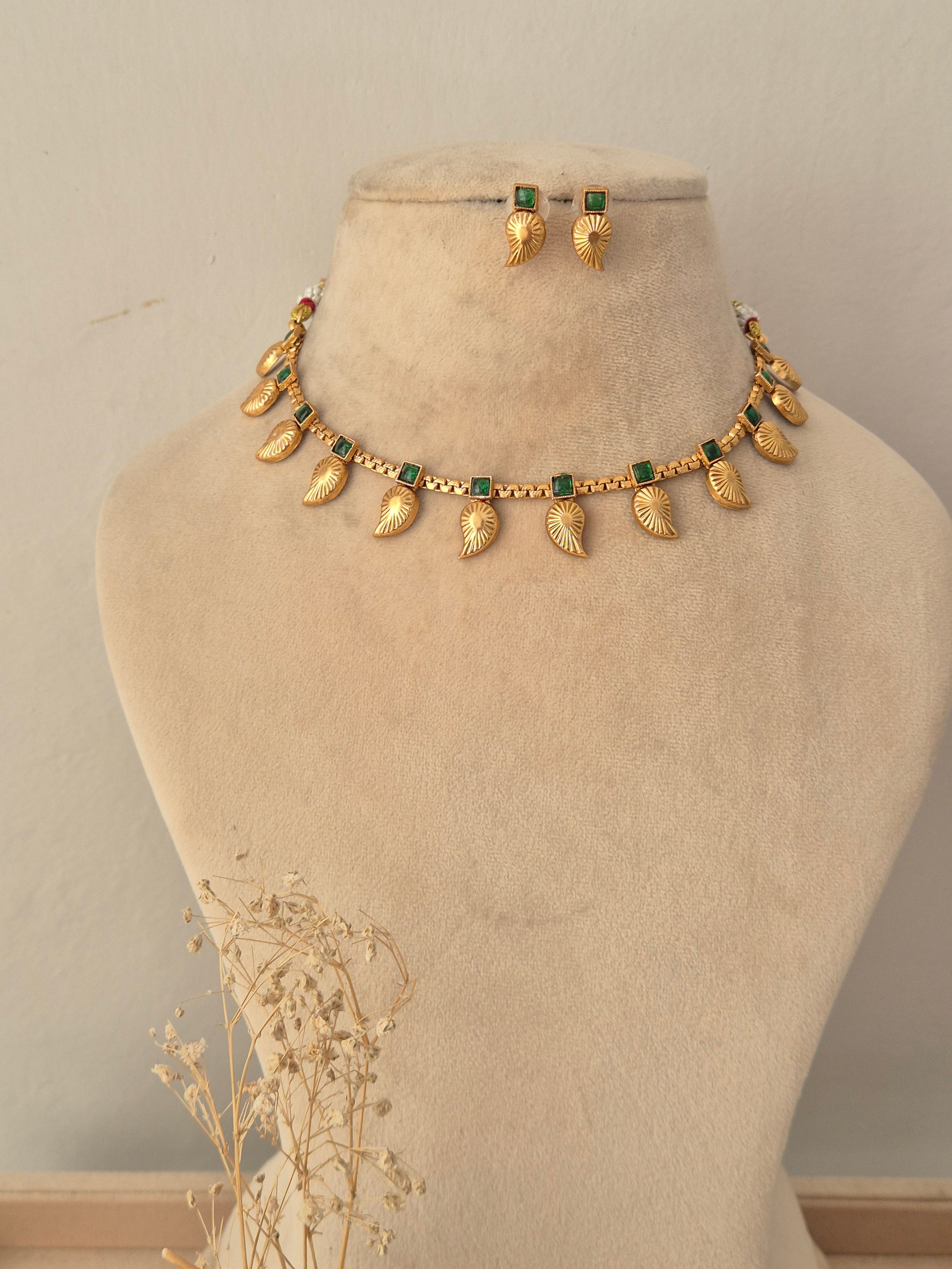 Tanvika Necklace Set showcasing an antique gold-toned choker embellished with rectangular green gemstone accents, delicate bead detailing, and hanging leaf-shaped charms, displayed on a beige mannequin bust alongside matching leaf drop earrings