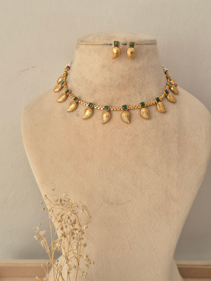 Tanvika Necklace Set showcasing an antique gold-toned choker embellished with rectangular green gemstone accents, delicate bead detailing, and hanging leaf-shaped charms, displayed on a beige mannequin bust alongside matching leaf drop earrings