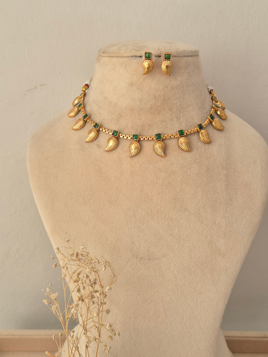 Tanvika Necklace Set showcasing an antique gold-toned choker embellished with rectangular green gemstone accents, delicate bead detailing, and hanging leaf-shaped charms, displayed on a beige mannequin bust alongside matching leaf drop earrings