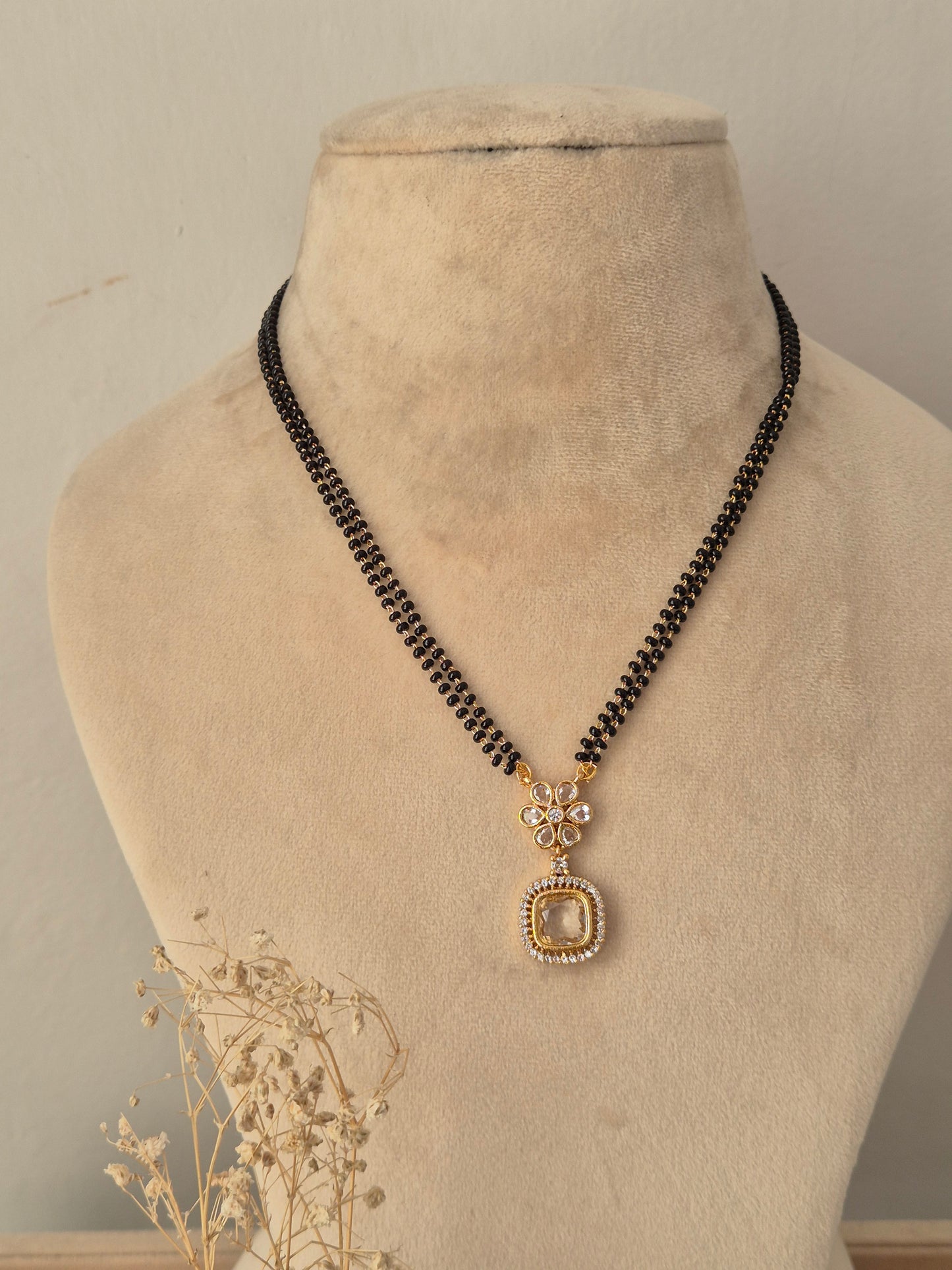 Kavika Mangalsutra featuring a traditional double-strand black and gold bead chain paired with a central gold-toned floral pendant studded with clear CZ stones and finished with a hanging teardrop accent