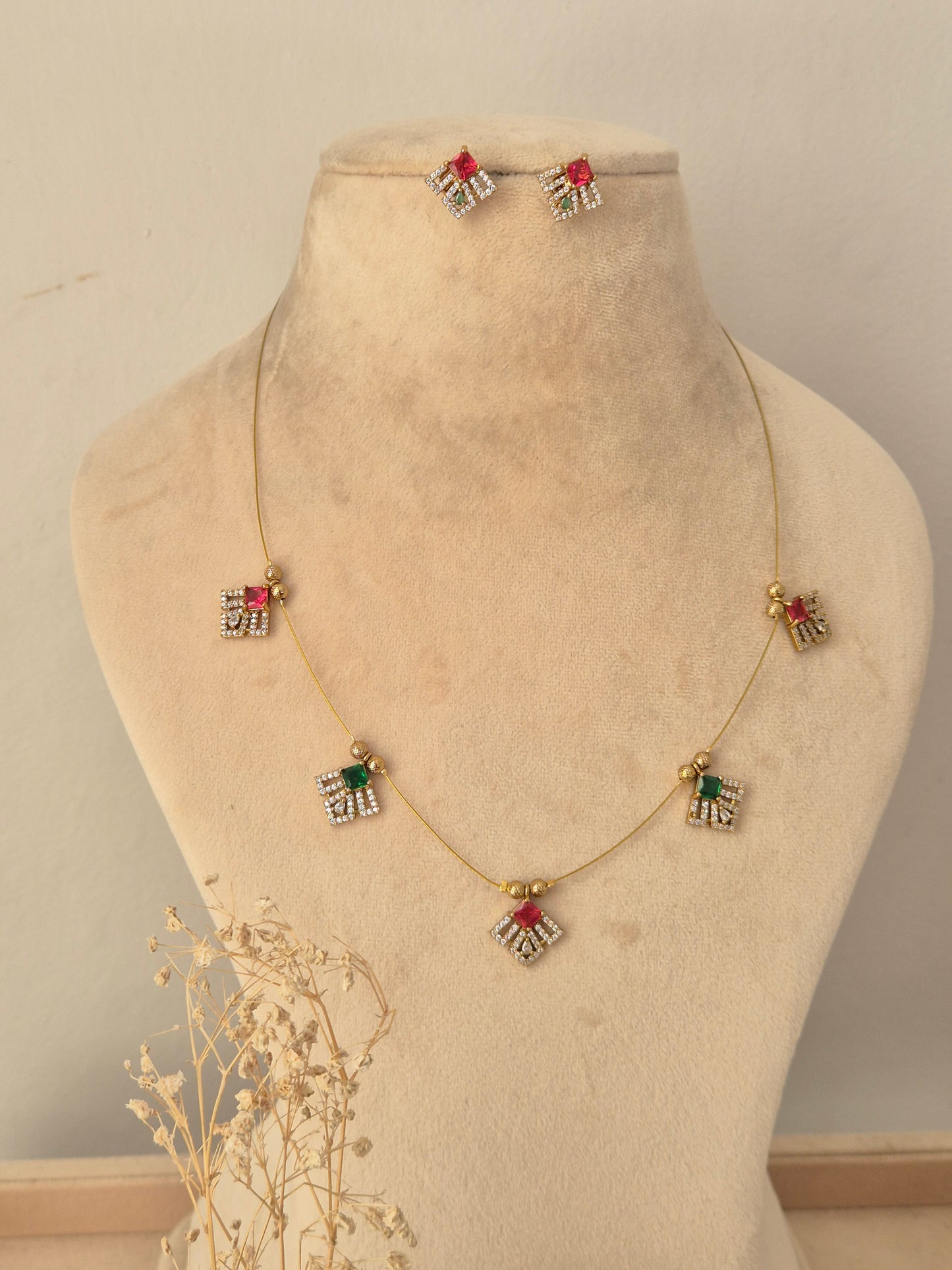 Labdhi Invisible Necklace Chain Set showcasing a fine gold-toned wire chain adorned with five geometric charms featuring red, green, and clear CZ stones, displayed on a beige mannequin bust alongside matching stud earrings.