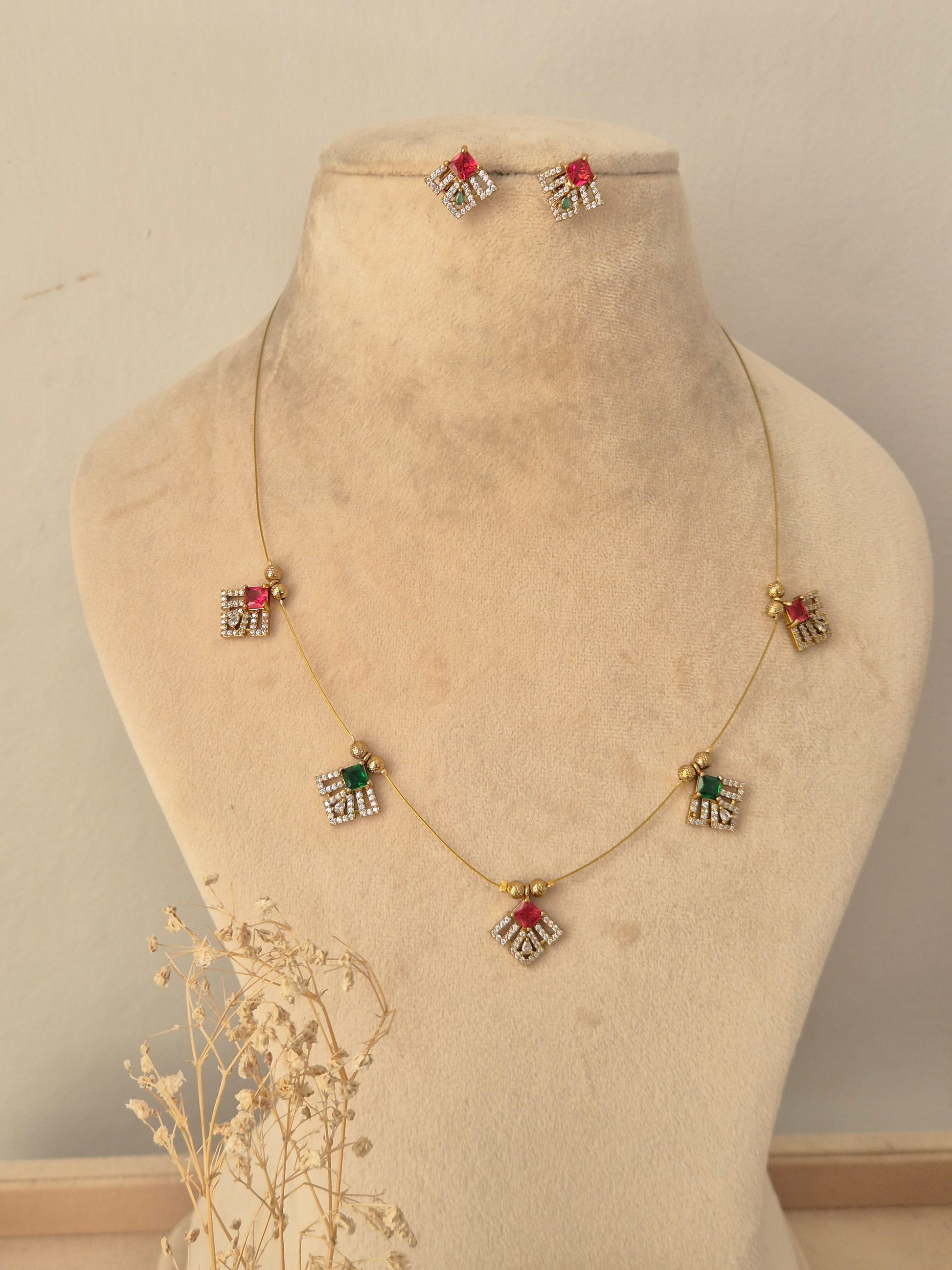 Labdhi Invisible Necklace Chain Set showcasing a fine gold-toned wire chain adorned with five geometric charms featuring red, green, and clear CZ stones, displayed on a beige mannequin bust alongside matching stud earrings.