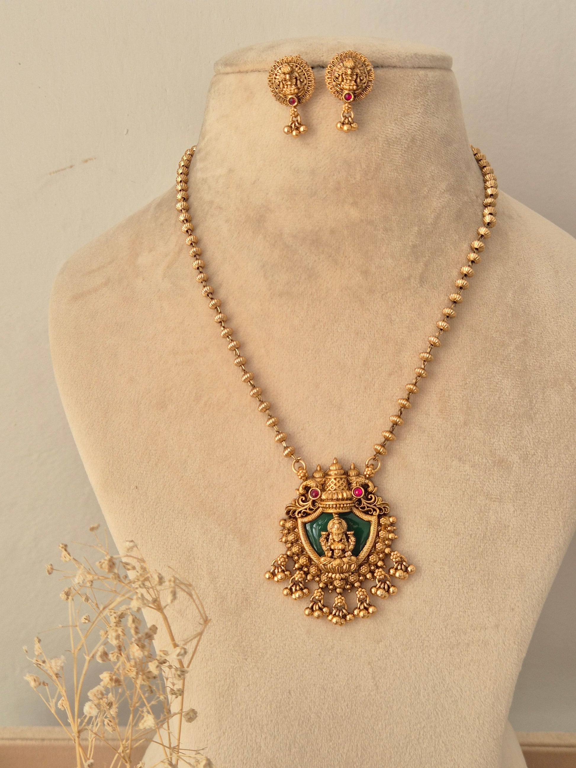Charumati Necklace Set featuring an antique gold-toned beaded chain holding a traditional temple pendant centered with a Goddess Lakshmi motif on a green enamel backdrop, accented by pink CZ stones and dangling gold cluster beads, displayed on a beige mannequin bust with matching round studs