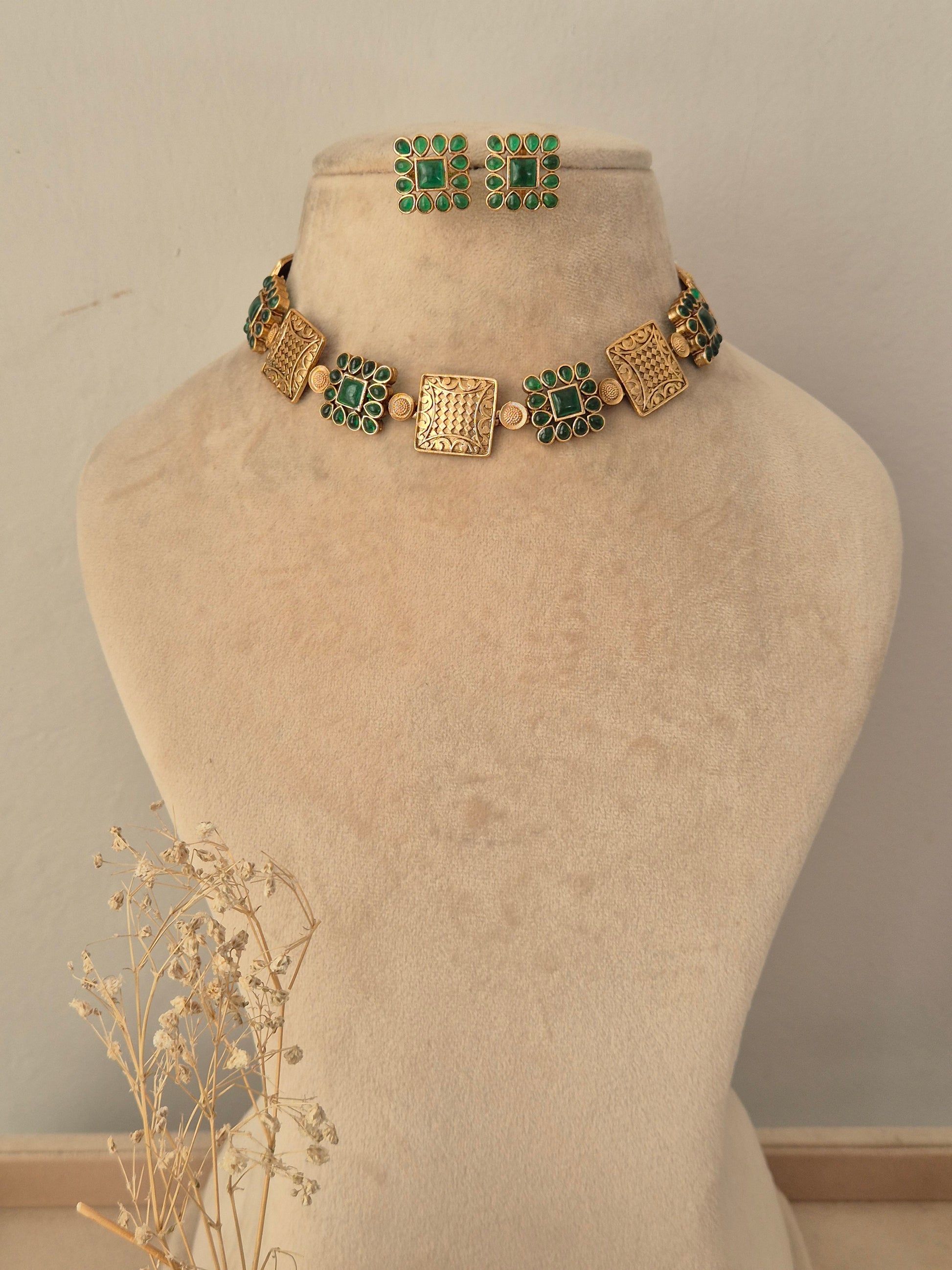 Lambika Necklace Set showcasing an antique gold-toned choker adorned with alternating square metal motifs and cluster-set green CZ stone accents, displayed on a beige mannequin bust with matching square studs