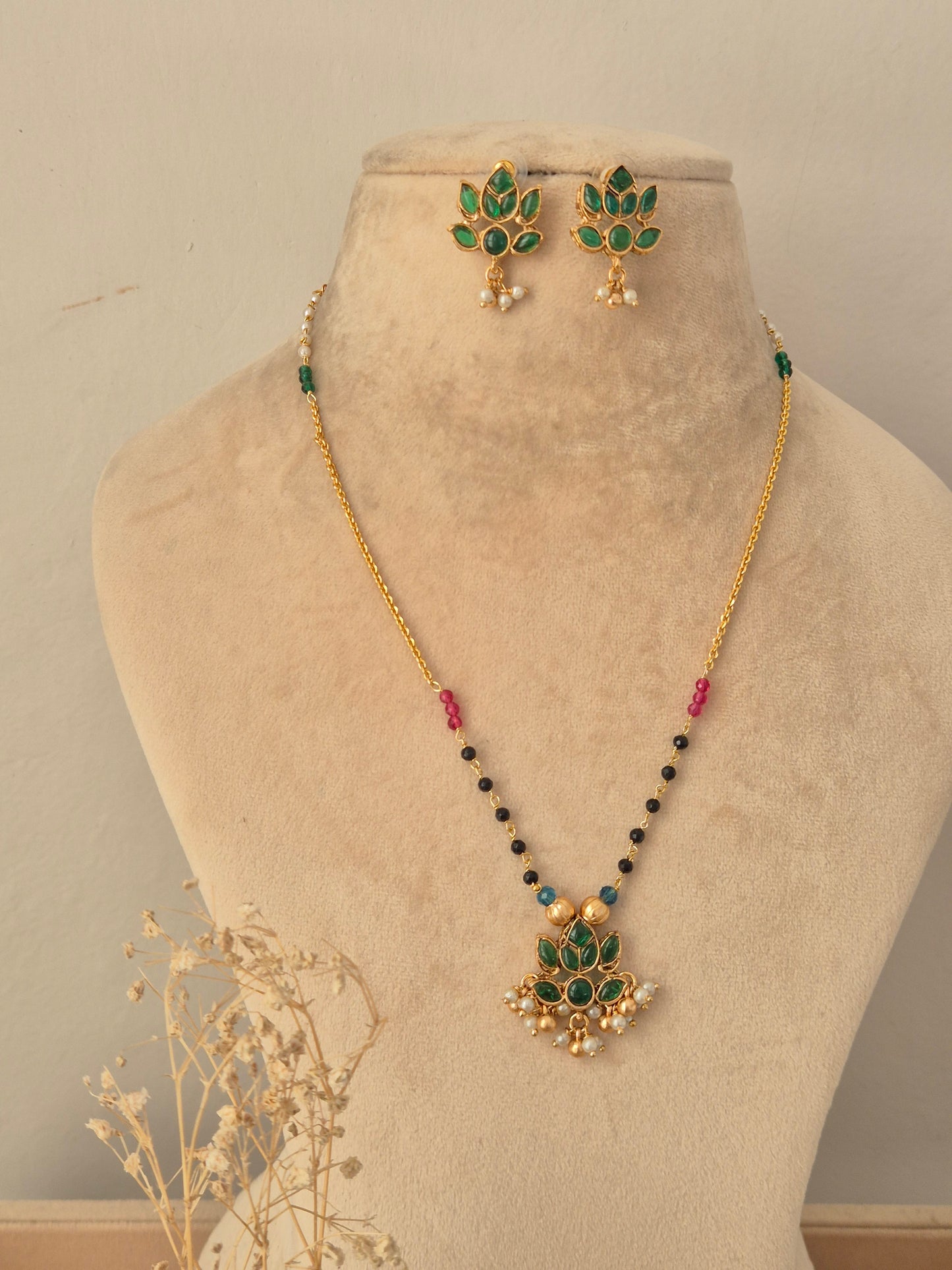 Prabhati Necklace Set showcasing a gold-toned chain with colorful bead stations and a green stone lotus-shaped pendant with pearl drops, displayed on a beige mannequin bust with matching floral stud earrings