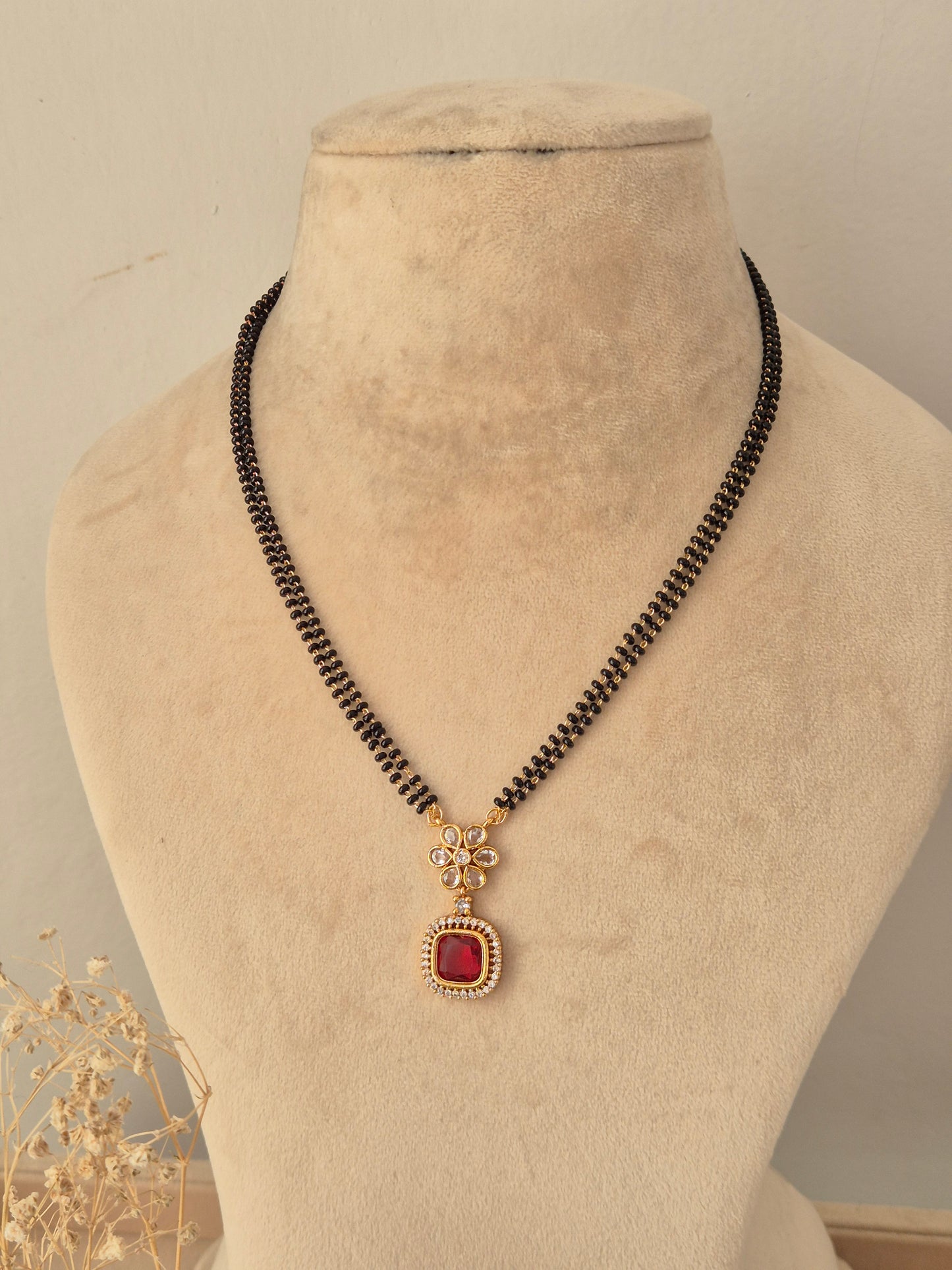 Prisham Mangalsutra featuring a double-strand black beaded chain with gold stations holding a gold floral pendant topped with a square red stone framed by CZ accents, displayed on a beige mannequin bust