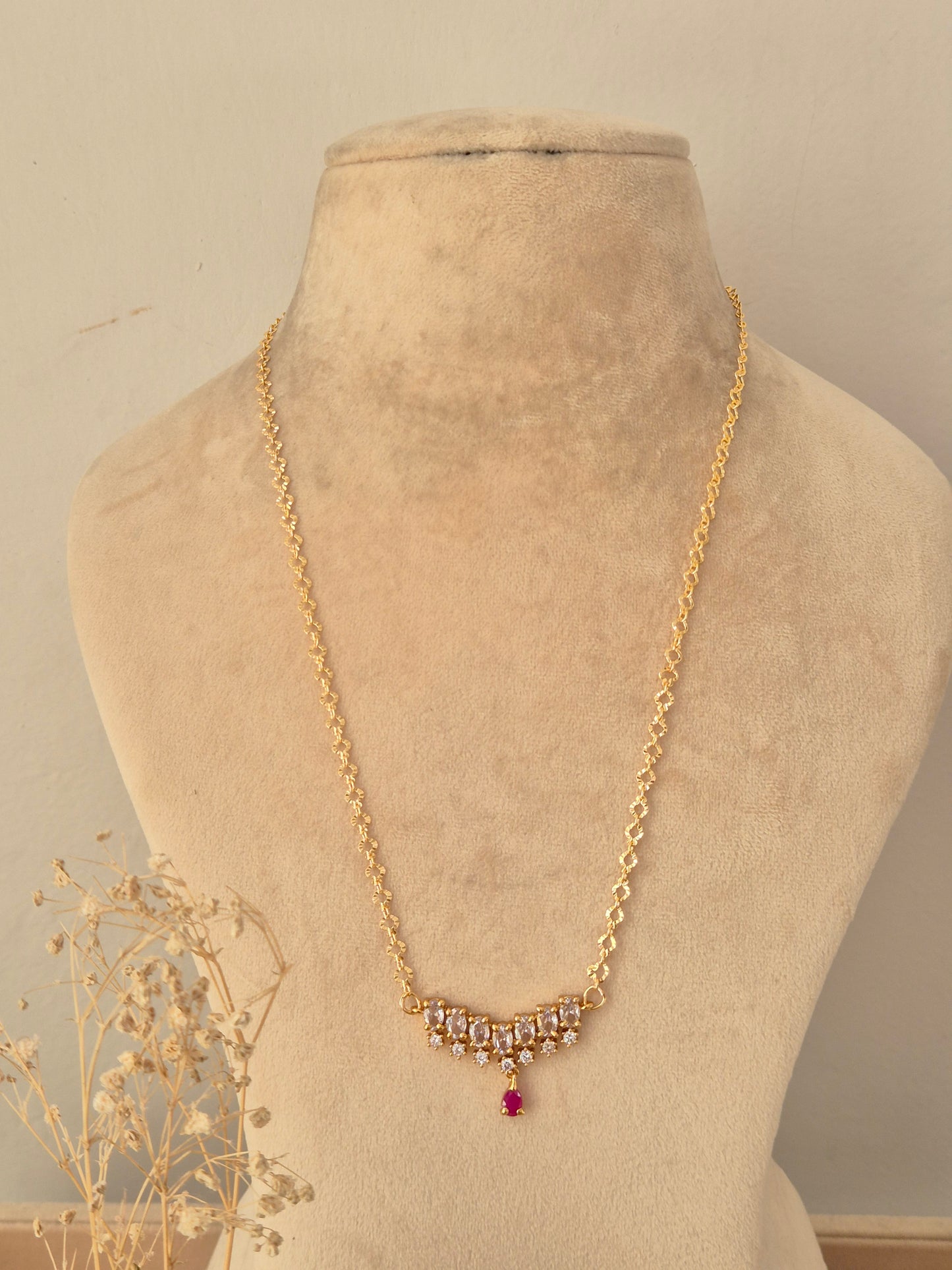 Zaahra Necklace showcasing a delicate gold-toned chain with a center arch of clear marquise CZ stones, anchored by a vibrant pinkish-red teardrop pendant drop, displayed on a beige mannequin bust