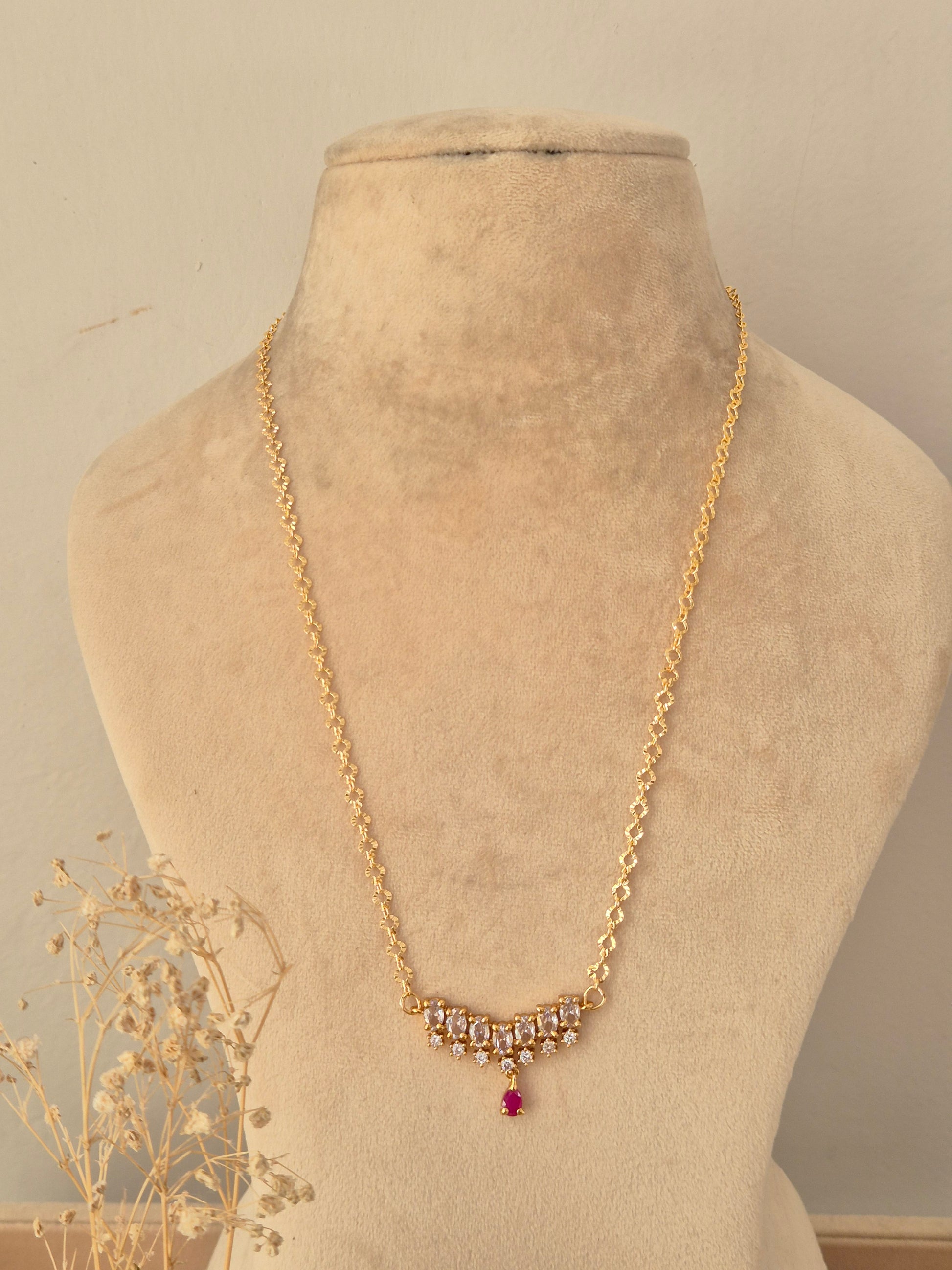 Zaahra Necklace showcasing a delicate gold-toned chain with a center arch of clear marquise CZ stones, anchored by a vibrant pinkish-red teardrop pendant drop, displayed on a beige mannequin bust
