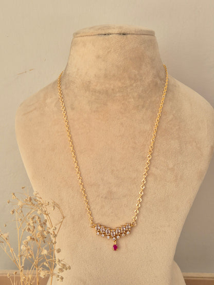 Zaahra Necklace showcasing a delicate gold-toned chain with a center arch of clear marquise CZ stones, anchored by a vibrant pinkish-red teardrop pendant drop, displayed on a beige mannequin bust