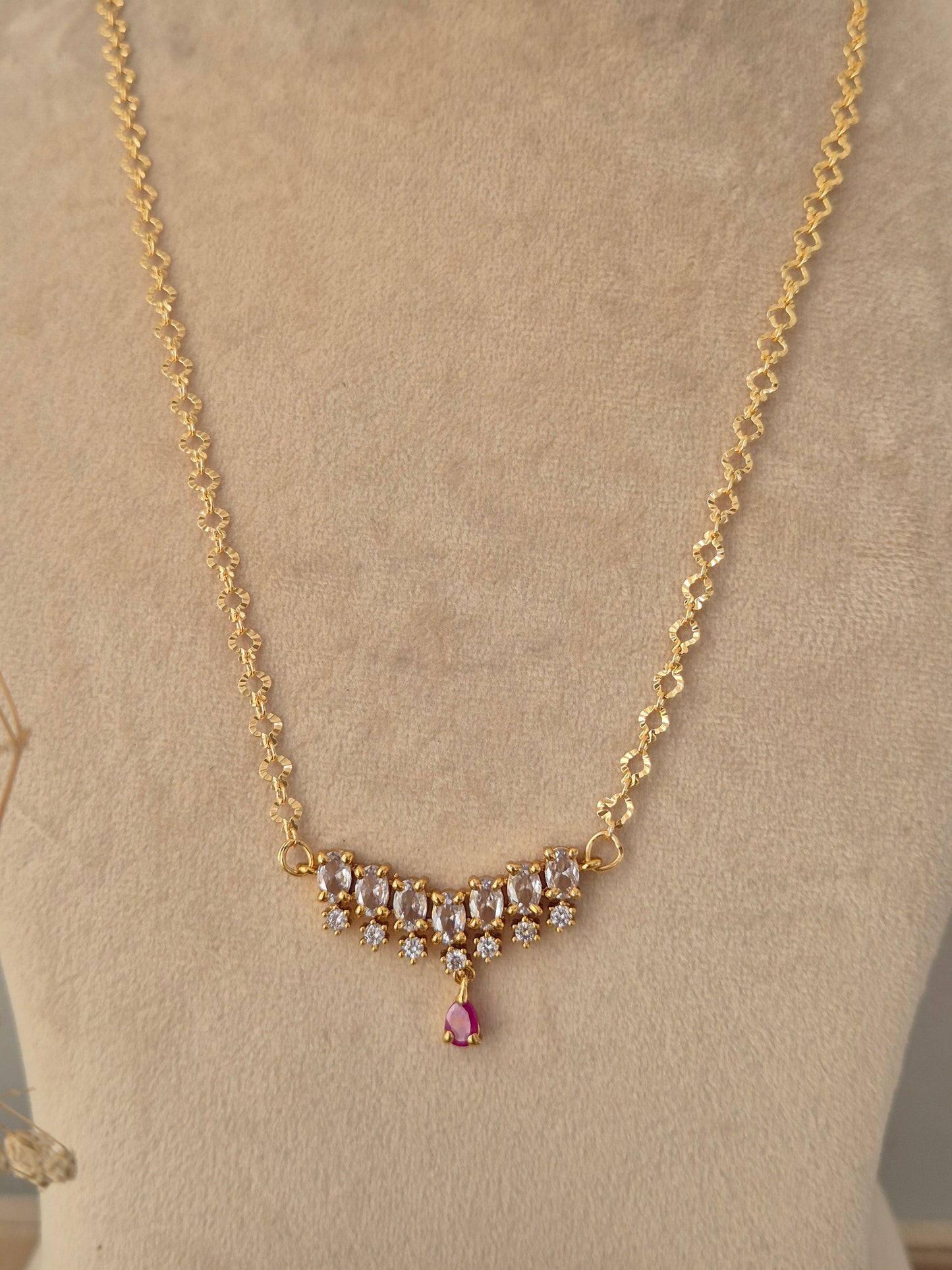 Zaahra Necklace showcasing a delicate gold-toned chain with a center arch of clear marquise CZ stones, anchored by a vibrant pinkish-red teardrop pendant drop, displayed on a beige mannequin bust