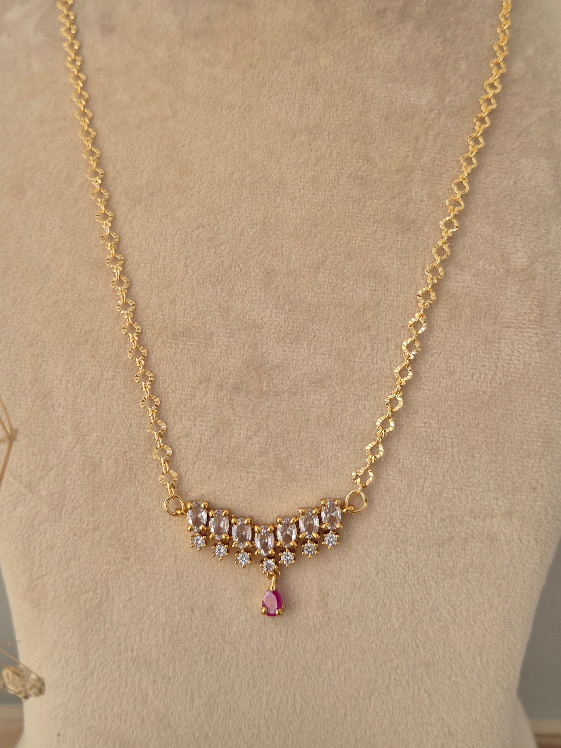 Zaahra Necklace showcasing a delicate gold-toned chain with a center arch of clear marquise CZ stones, anchored by a vibrant pinkish-red teardrop pendant drop, displayed on a beige mannequin bust