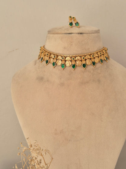 Traditional temple jewellery choker set featuring gold Laxmi coin motifs lined with green stone drop pendants, paired with matching small green-stone earrings on a neutral display mannequin