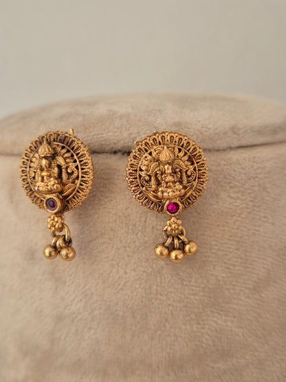 Pair of round antique gold temple earrings featuring a central Goddess motif, ruby-pink stone accent, and dangling gold beads on a beige stand