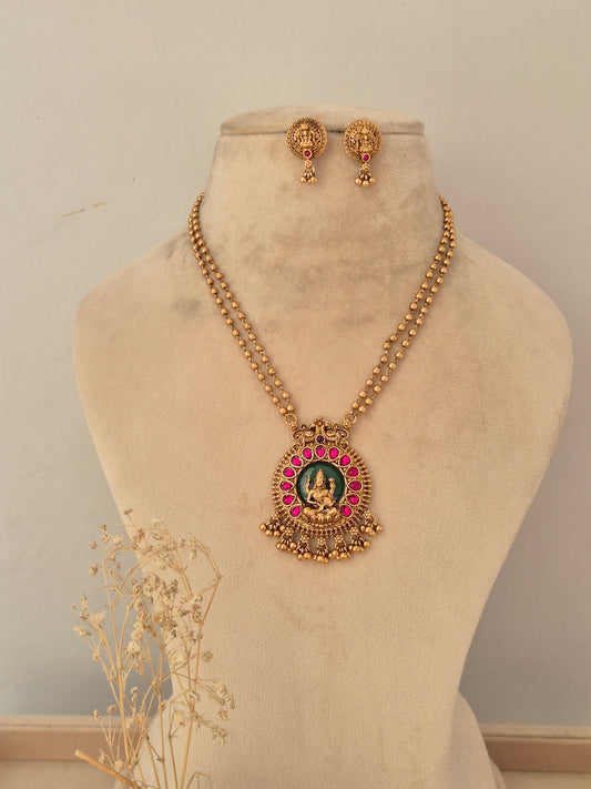 Antique gold long temple necklace set featuring a circular Goddess Laxmi pendant set against a green background, encircled by ruby-pink stones, with matching stud earrings