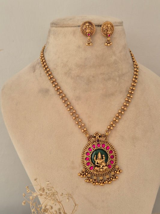 Traditional Indian long-chain temple jewellery set showcasing a double beaded chain and a circular central Goddess Laxmi motif pendant accented with emerald-green inlay, framed by a halo of ruby-pink stones and gold drop beads, complete with matching deity stud earrings