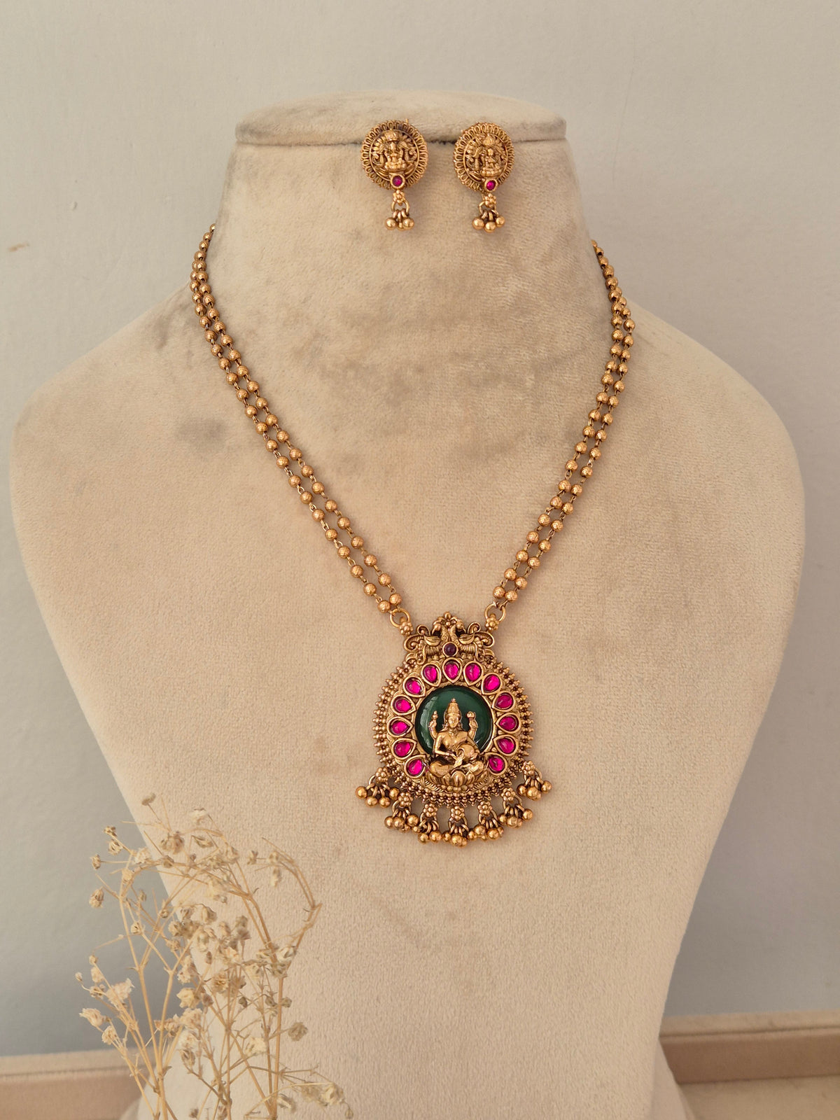 Traditional long antique gold temple necklace featuring a green and ruby-pink Laxmi pendant and matching stud earrings on a display bust