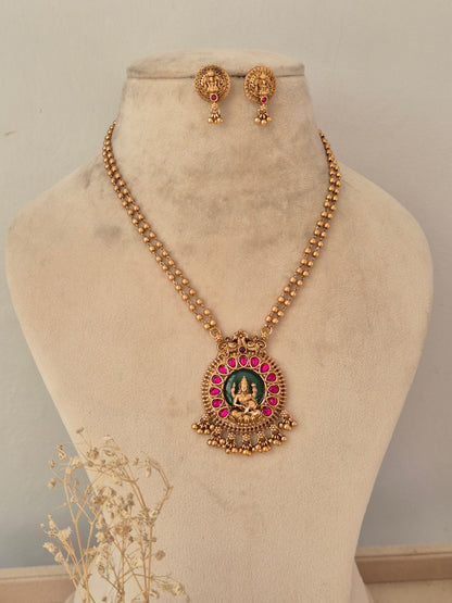 Traditional long antique gold temple necklace featuring a green and ruby-pink Laxmi pendant and matching stud earrings on a display bust