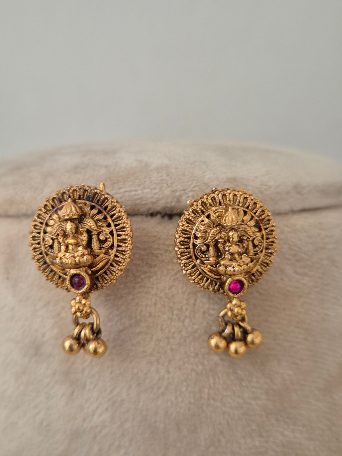 Option 1 (Short): Pair of circular antique gold temple earrings featuring a central Goddess Laxmi motif with a ruby-pink stone drop and gold bead dangles