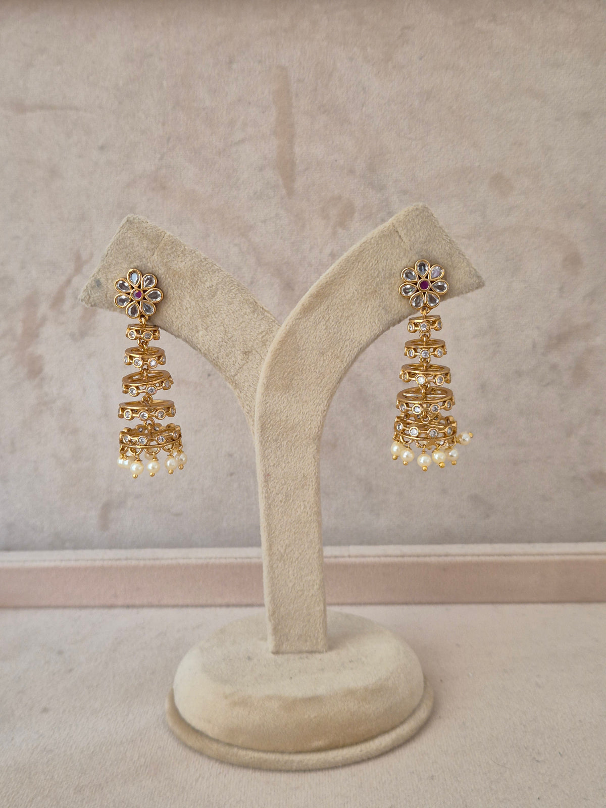 A pair of gold-toned tiered jhumka earrings with floral stone-studded tops and pearl dangles, displayed on a beige velvet jewelry stand