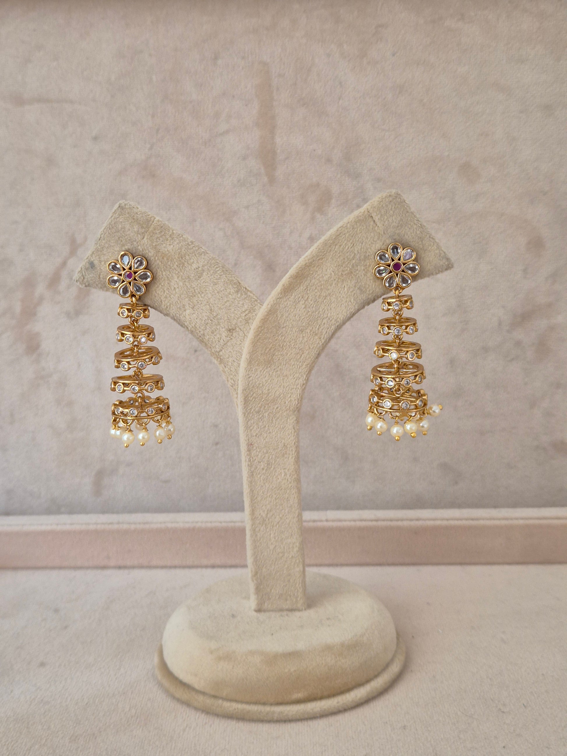 A pair of gold-toned tiered jhumka earrings with floral stone-studded tops and pearl dangles, displayed on a beige velvet jewelry stand