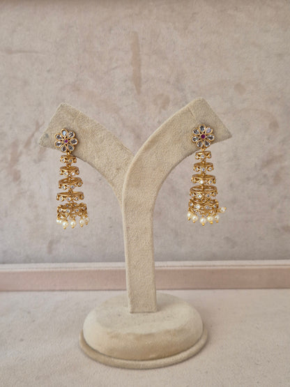 A pair of gold-toned tiered jhumka earrings with floral stone-studded tops and pearl dangles, displayed on a beige velvet jewelry stand