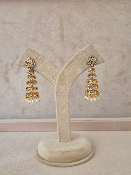A pair of gold-toned tiered jhumka earrings with floral stone-studded tops and pearl dangles, displayed on a beige velvet jewelry stand