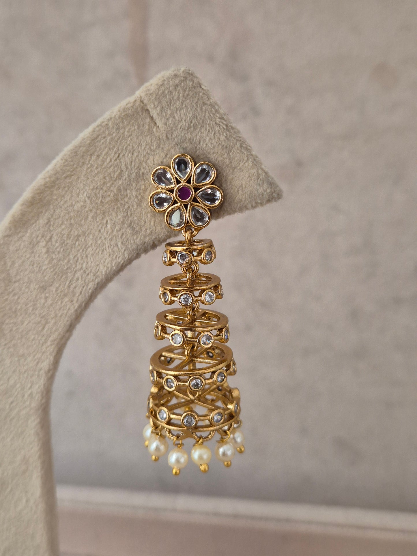 Traditional Indian gold-finish multi-tiered jhumka earring featuring a flower-shaped stud with a central purple-ruby stone, stone-studded wireframe bells, and small pearl bead drops