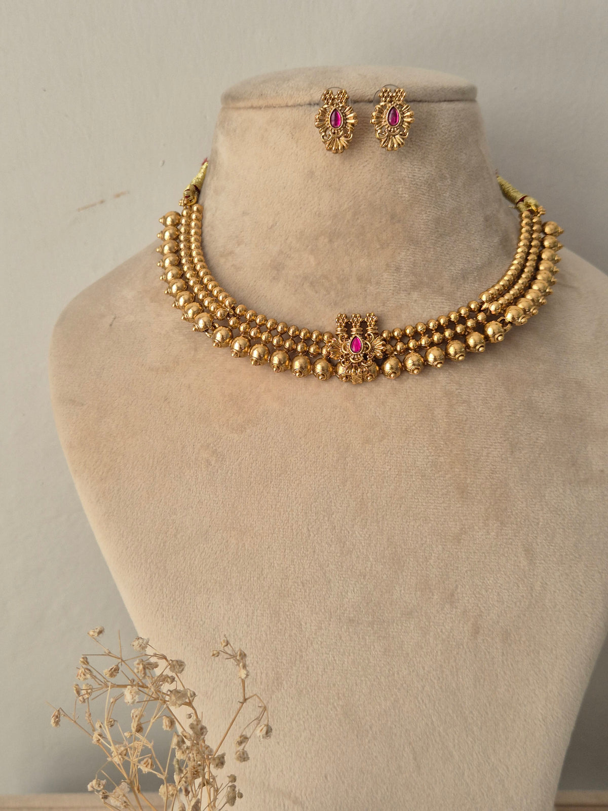 Traditional Indian short necklace set in antique gold finish, featuring three rows of polished gold beads connected to a center floral pendant set with a ruby-pink stone, complete with matching round stud earrings