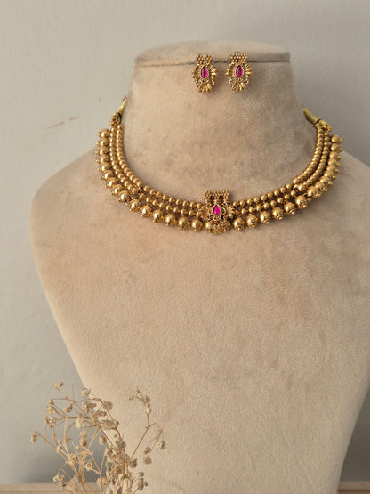 Traditional Indian short necklace set in antique gold finish, featuring three rows of polished gold beads connected to a center floral pendant set with a ruby-pink stone, complete with matching round stud earrings