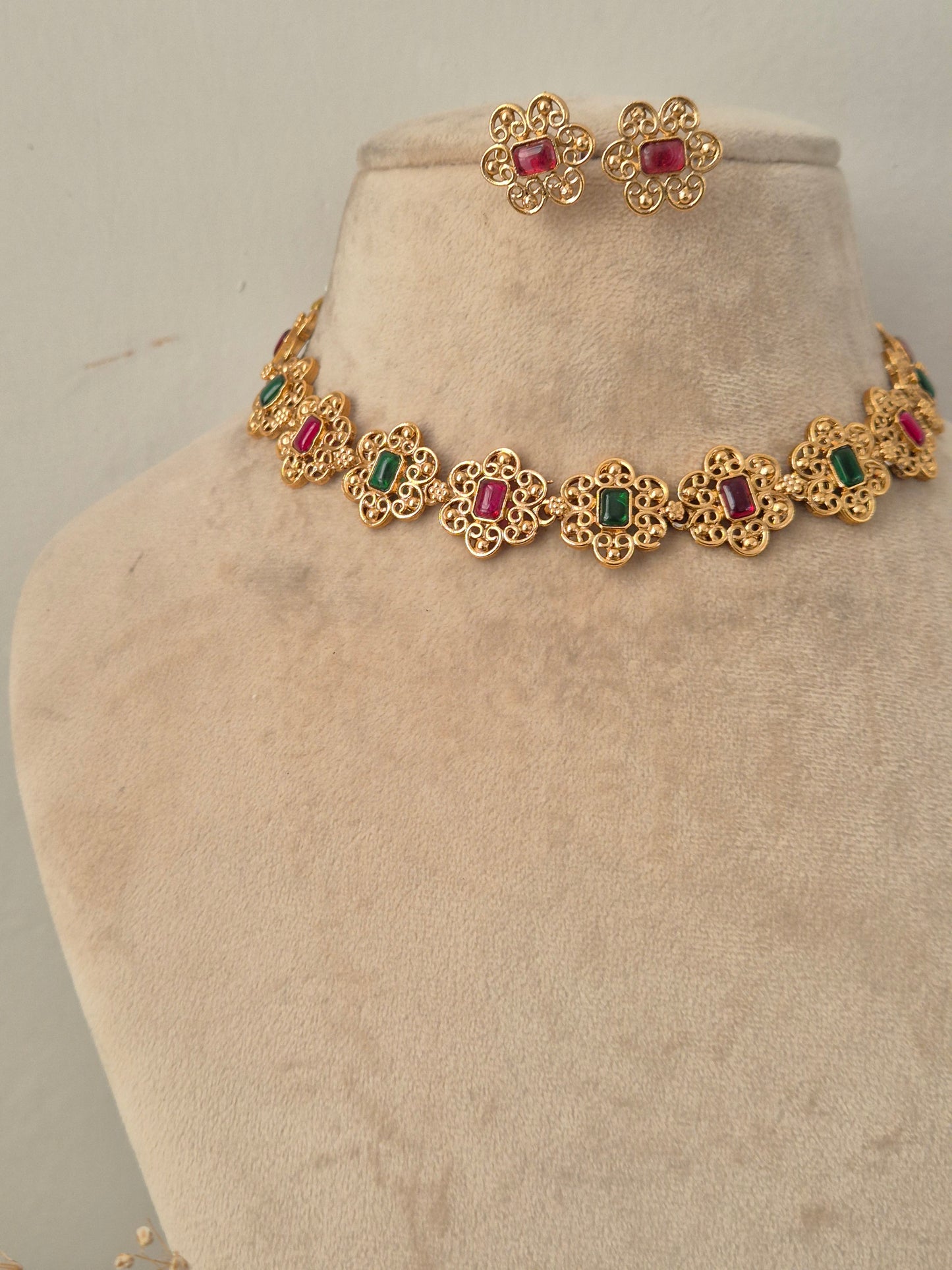 Traditional Indian antique gold-finish choker featuring connected floral motifs with alternating rectangular ruby-red and emerald-green gemstones, paired with matching floral stud earrings set with ruby-red stones