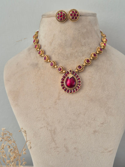 Traditional Indian antique gold necklace set featuring a chain studded with oval ruby-pink stones leading to a central teardrop ruby-pink pendant framed by smaller stones, paired with matching round floral stud earrings