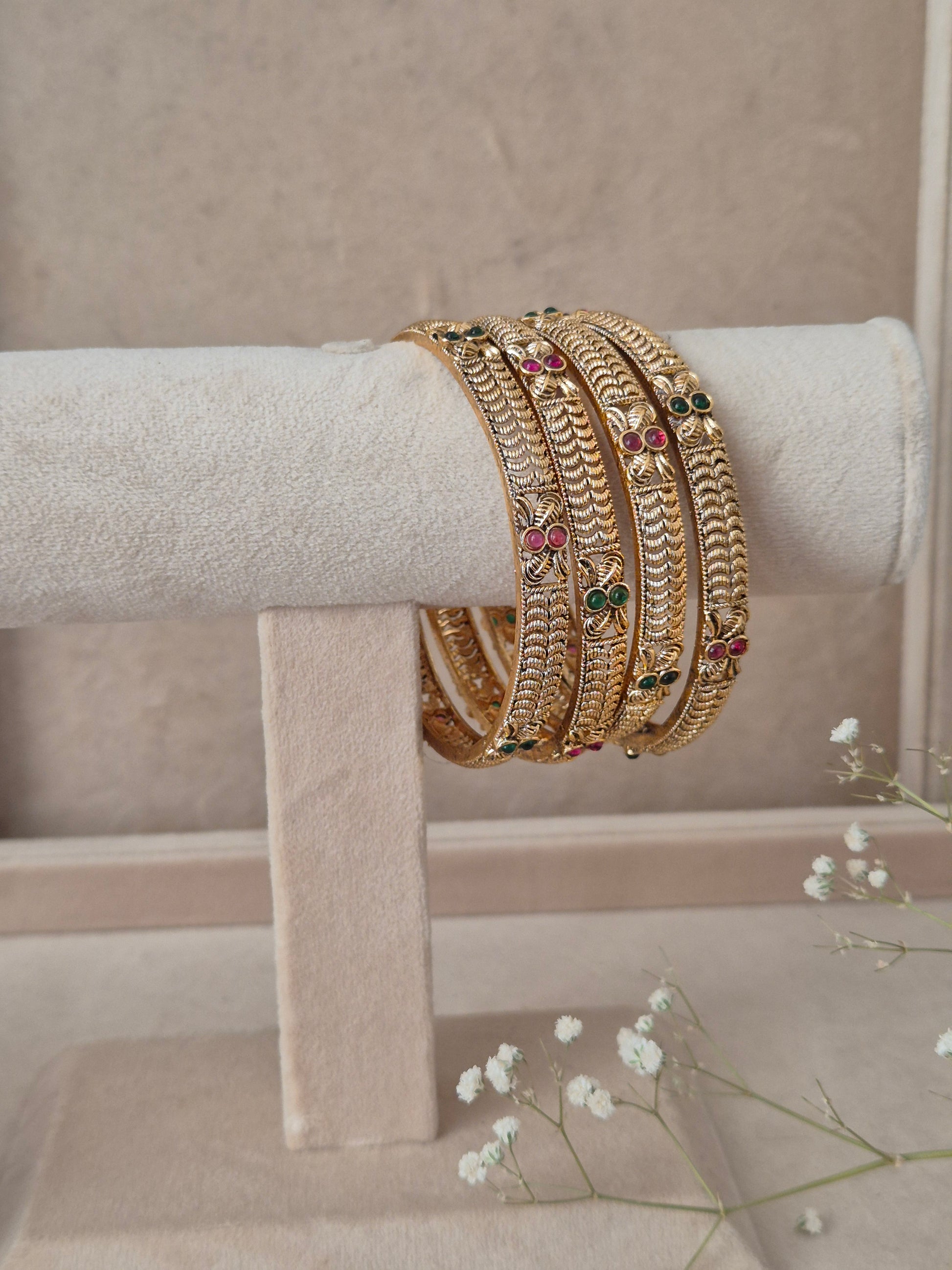 Four traditional gold-plated ethnic bangles with detailed floral patterns and inlaid red and green gemstones by naitri.in