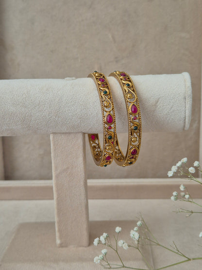 Two traditional gold bangles with intricate floral patterns, inlaid with red and green gemstones by naitri.in